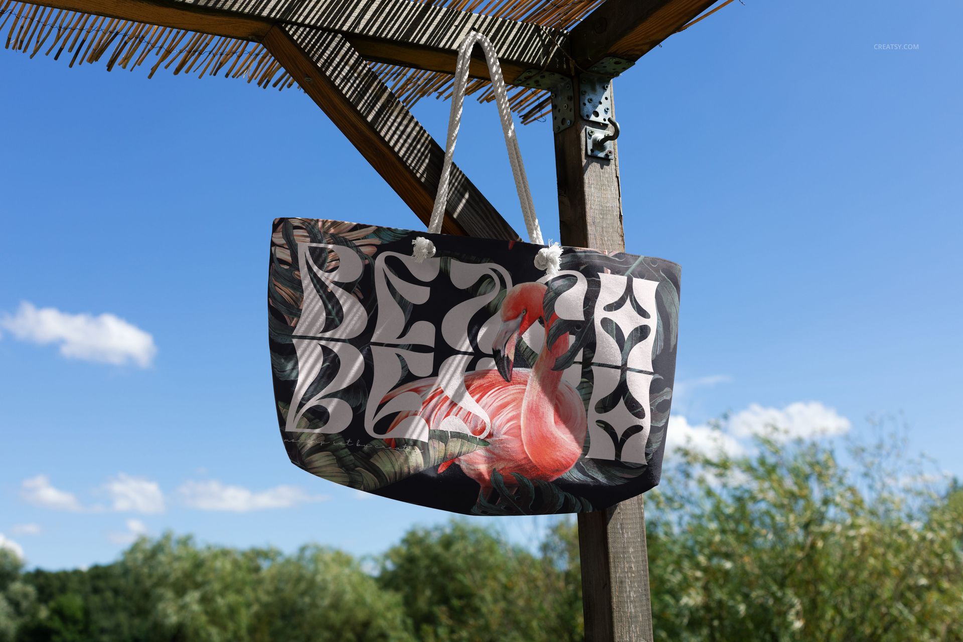 Weekender bag mockup hanging outdoors under a wooden pavilion, featuring a flamingo and leaf pattern on dark fabric.