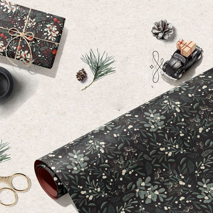 Gift wrapping paper mockup featuring a dark floral winter foliage pattern, rolled out on a textured surface with festive props.