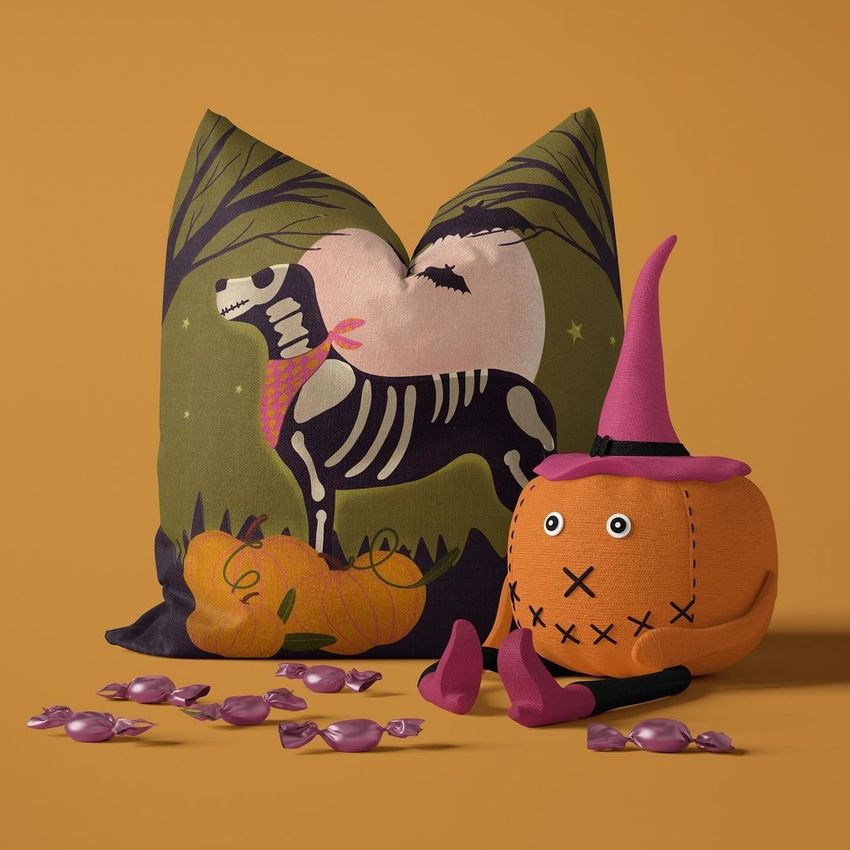 Halloween pillow mockup featuring a skeleton dog illustration next to a plush pumpkin toy with a witch hat on a brown surface.
