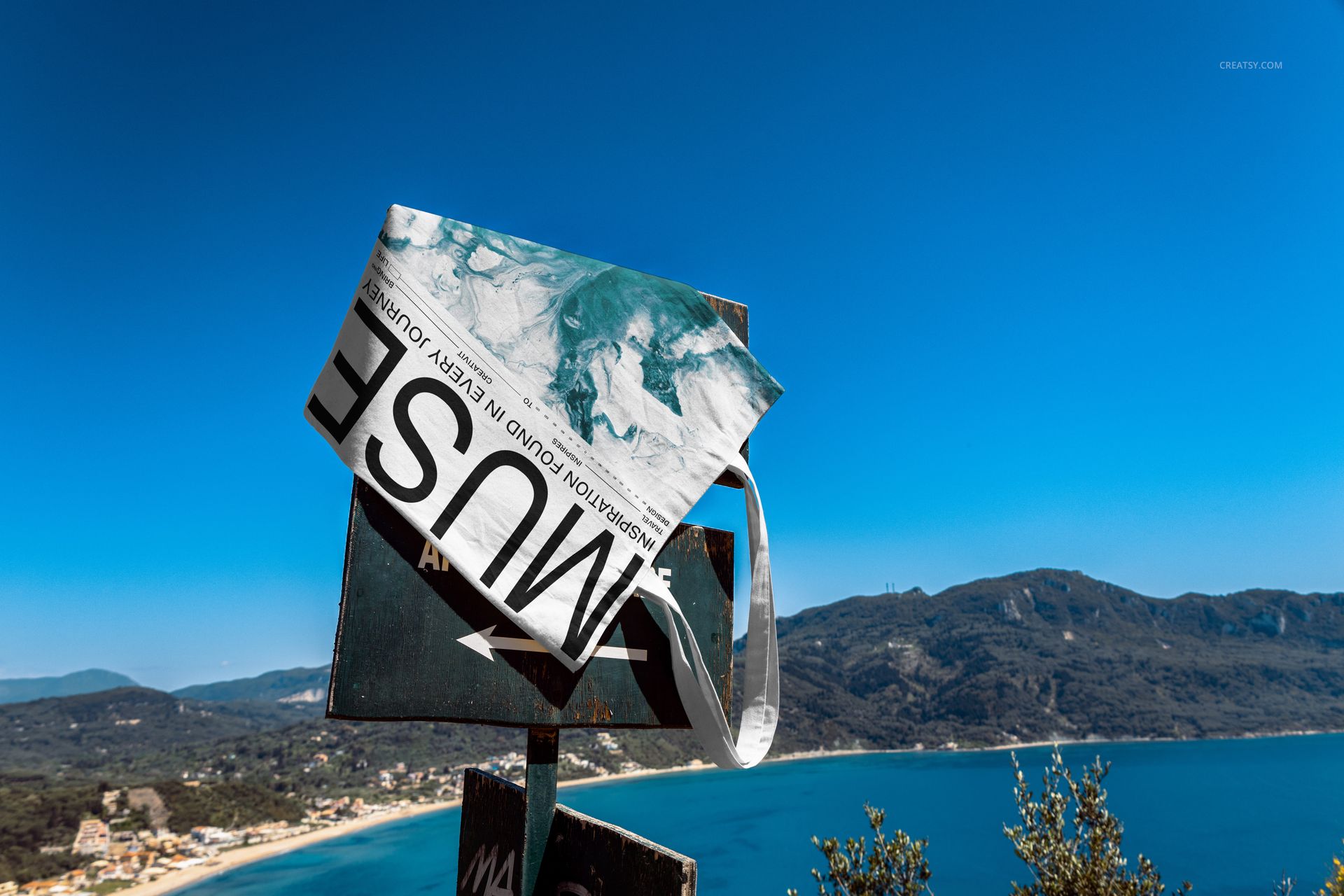 Outdoor scene featuring a tote bag mockup with bold text and blue pattern, set against sea and mountains background.