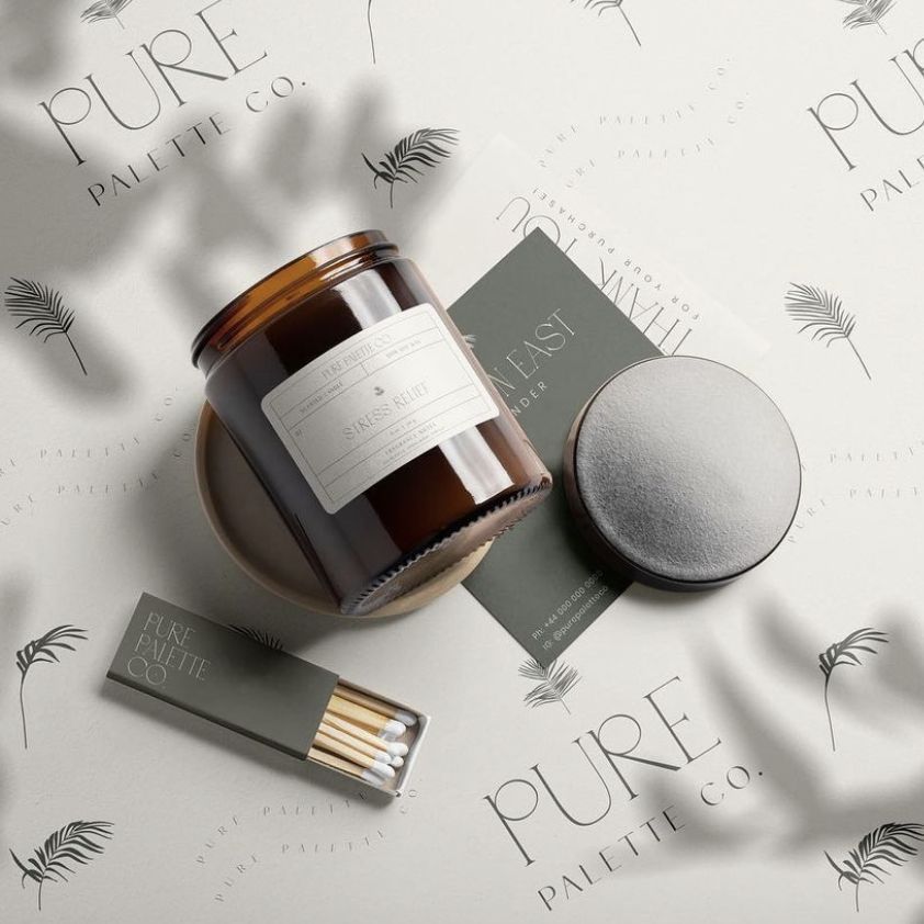 Sticker mockup scene with a brown glass candle jar, matchbox, and round sticker on a light background with leaf patterns.