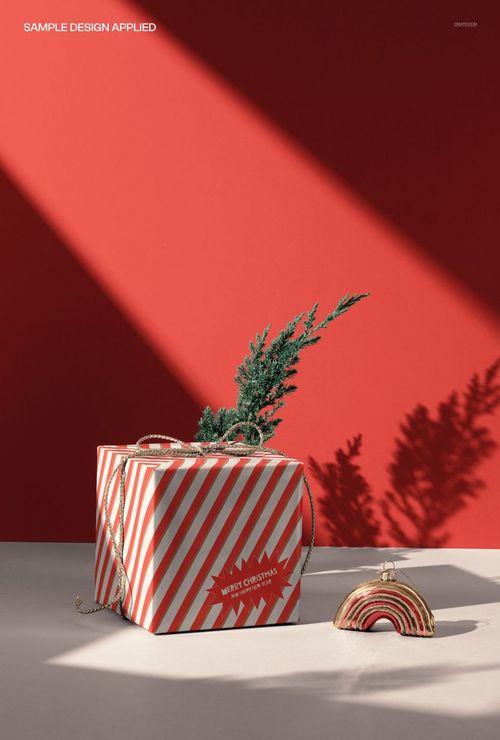 Christmas gift box mockup featuring striped red and white pattern, gold rope handles, and rainbow ornament in sunlight.