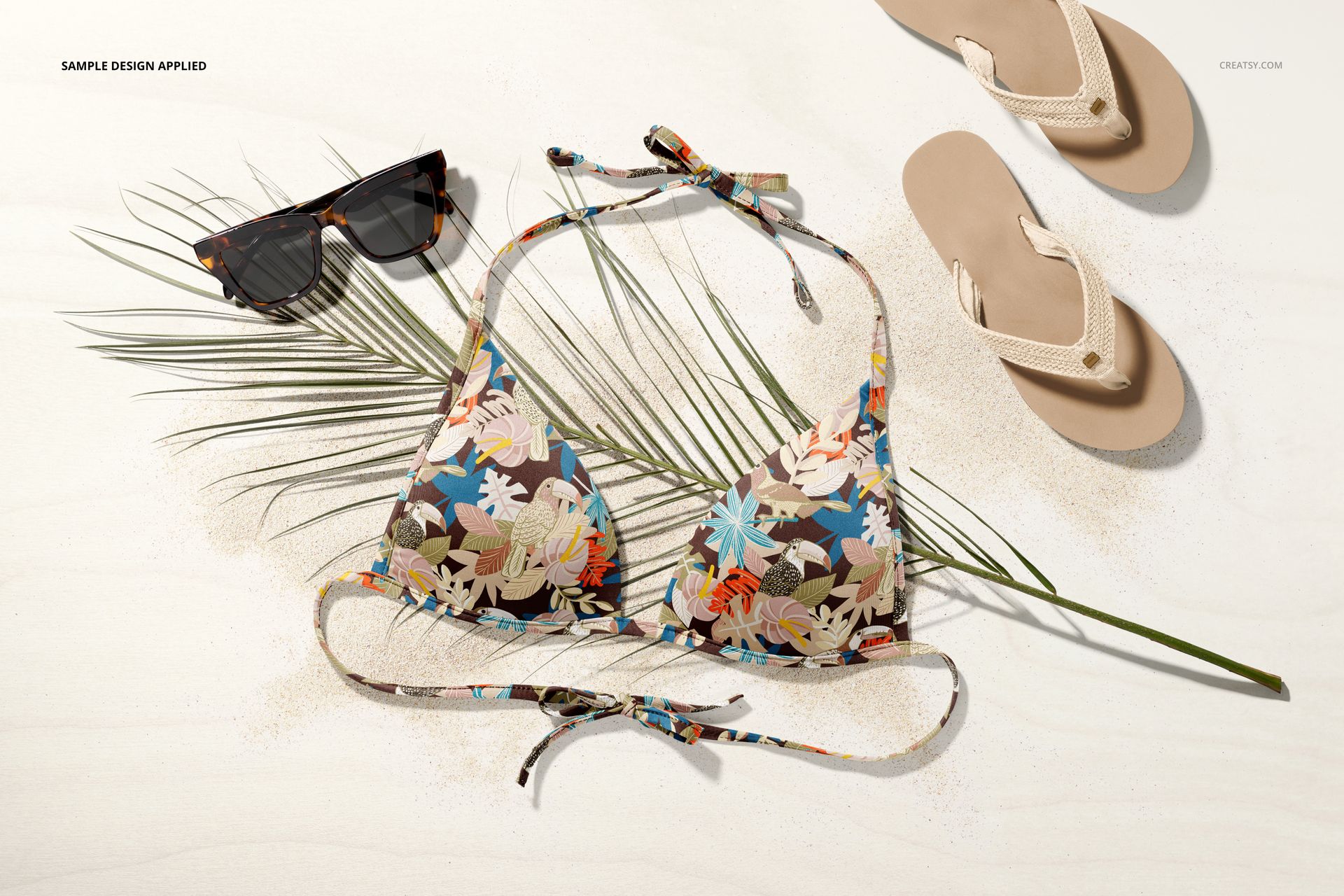 Floral patterned bikini top mockup displayed on a light surface with sunglasses, flip-flops, palm leaf, and sand.