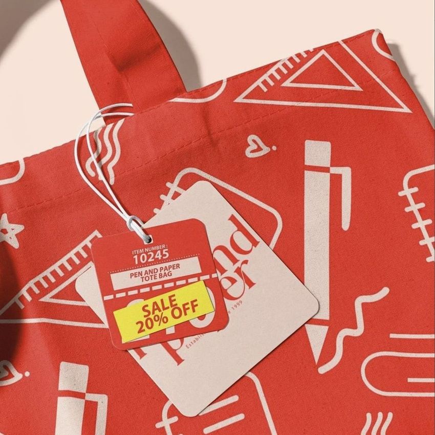 Canvas tote bag mockup featuring a red bag with white stationery-themed illustrations, close-up of handle and attached tags.