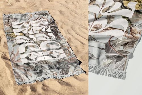 Fringed beach towel mockup featuring a white graphic over tropical leaf and animal print, shown on sand.