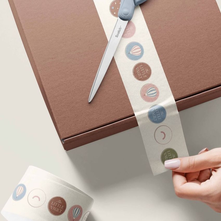 Mailing box mockup with a brown cardboard box on a light surface, decorated with a strip of patterned tape being cut by scissors.