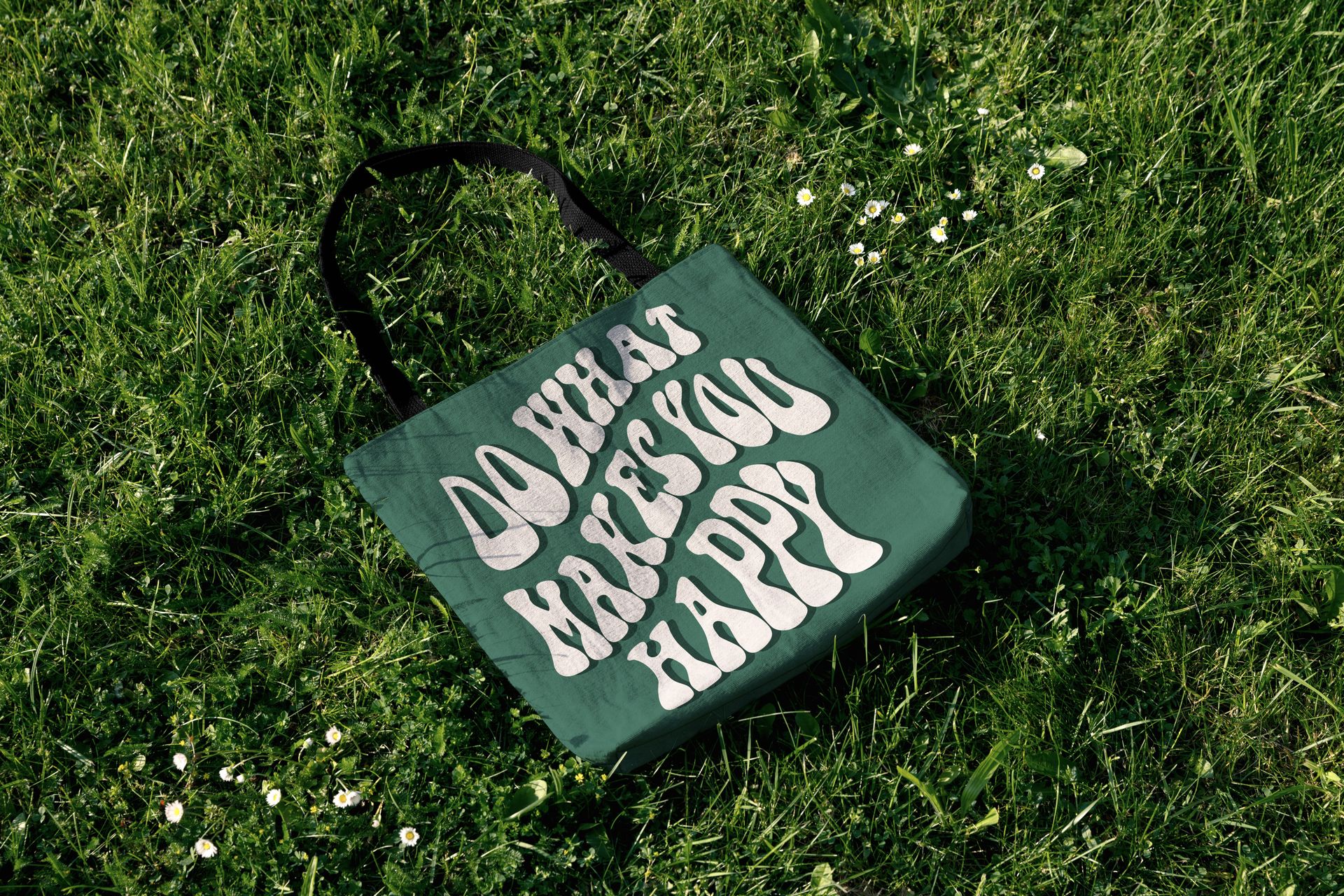 Outdoor mockup of a woven tote bag with dark handles and stylized white text, lying in a sunlit grassy area.