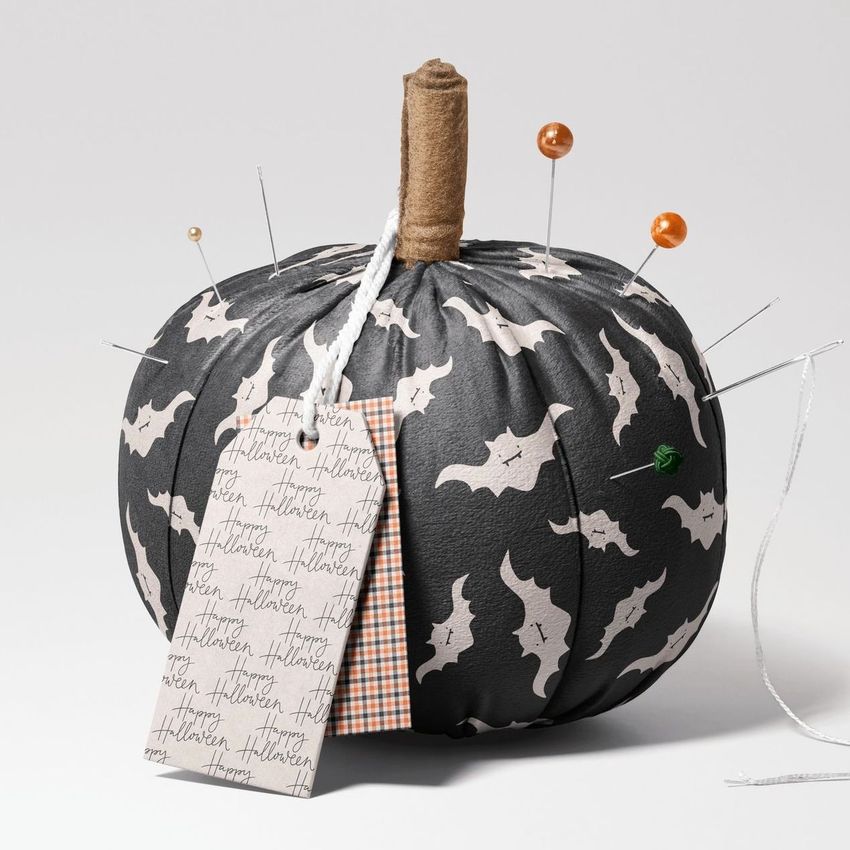 Fabric pumpkin mockup with a black surface decorated with white bat patterns, pins, a brown stem, and attached tags.