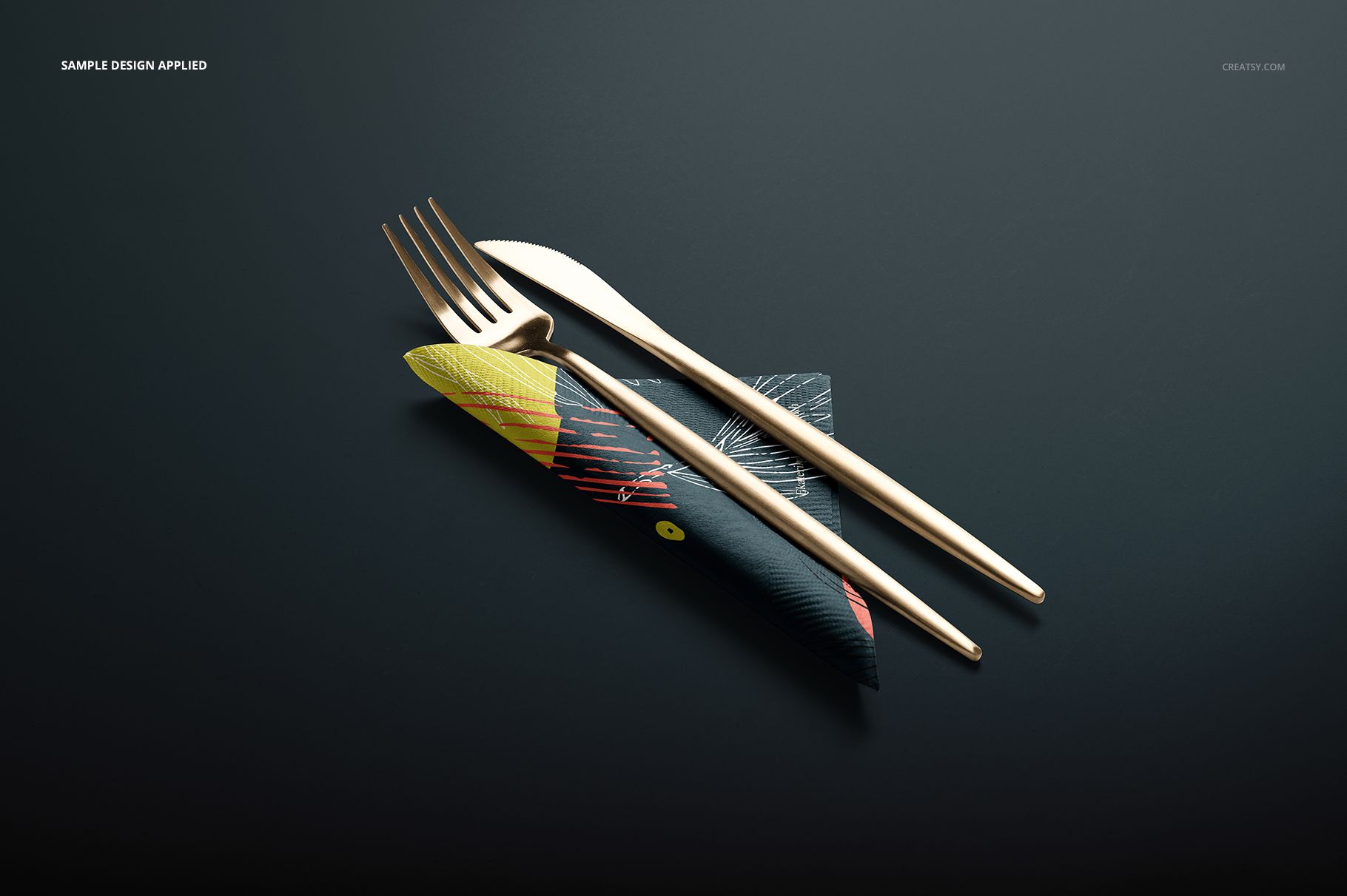 PSD mockup featuring a paper napkin with modern artwork, holding gold cutlery, arranged on a sleek dark tabletop.