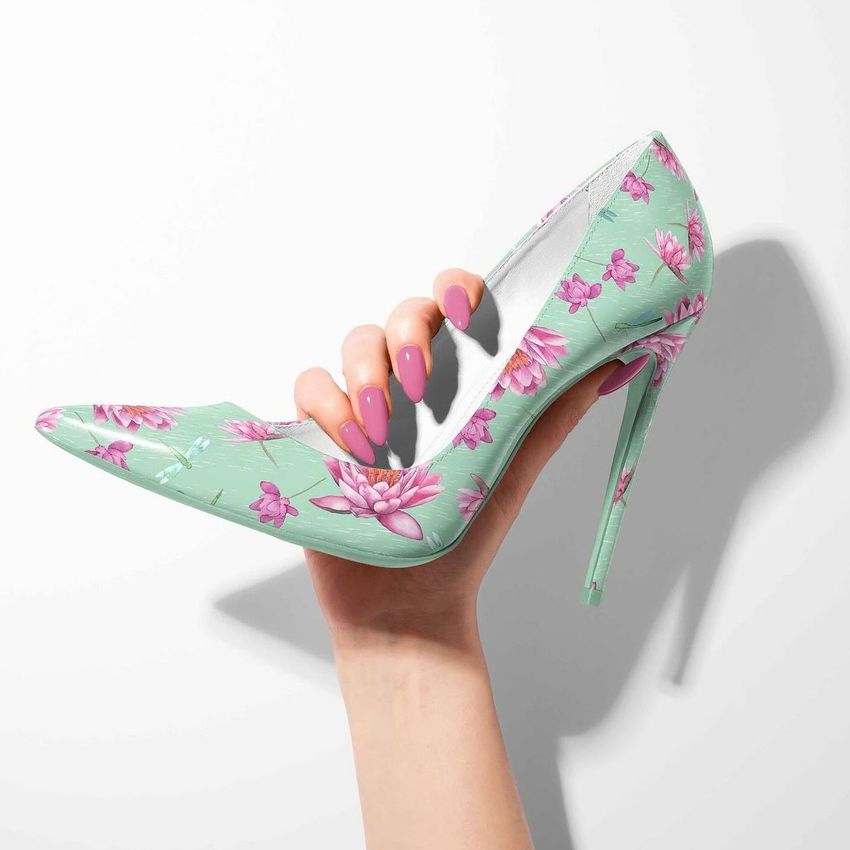 A hand with pink manicured nails holds a high heel mockup featuring a mint green base with pink waterlily and dragonfly patterns.