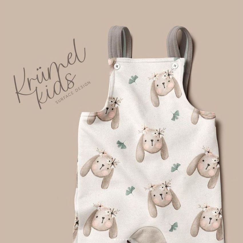 Kids fleece dungarees mockup featuring a cream fabric with a repeating bunny face pattern, shown on a beige background.