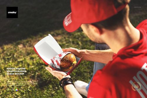 PSD mockup showing a hand holding a burger in a branded paper wrap inside a red and white box, outdoor setting.