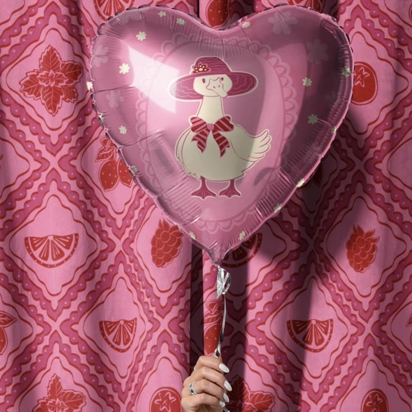 Hand holding a heart-shaped balloon with a duck illustration, set against a pink velvet curtain with fruit patterns.