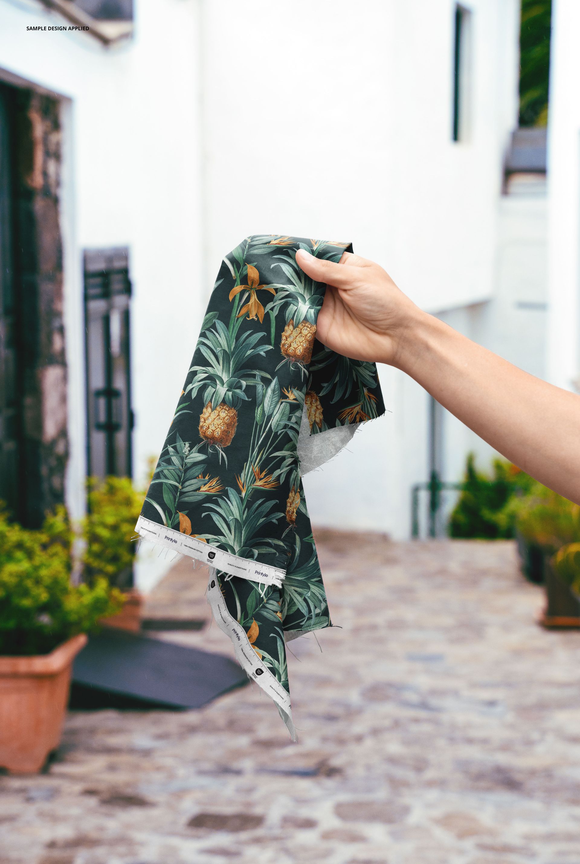 Hand holding a fabric sample with a tropical pineapple pattern against a rustic urban outdoor backdrop.