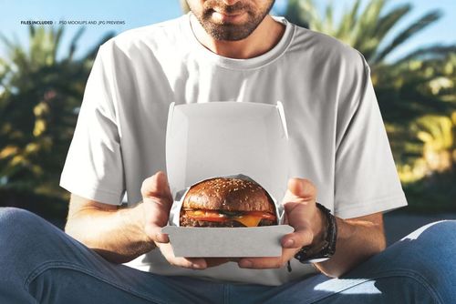 Close-up of a person in a white t-shirt presenting a burger box mockup, featuring a visible cheeseburger.