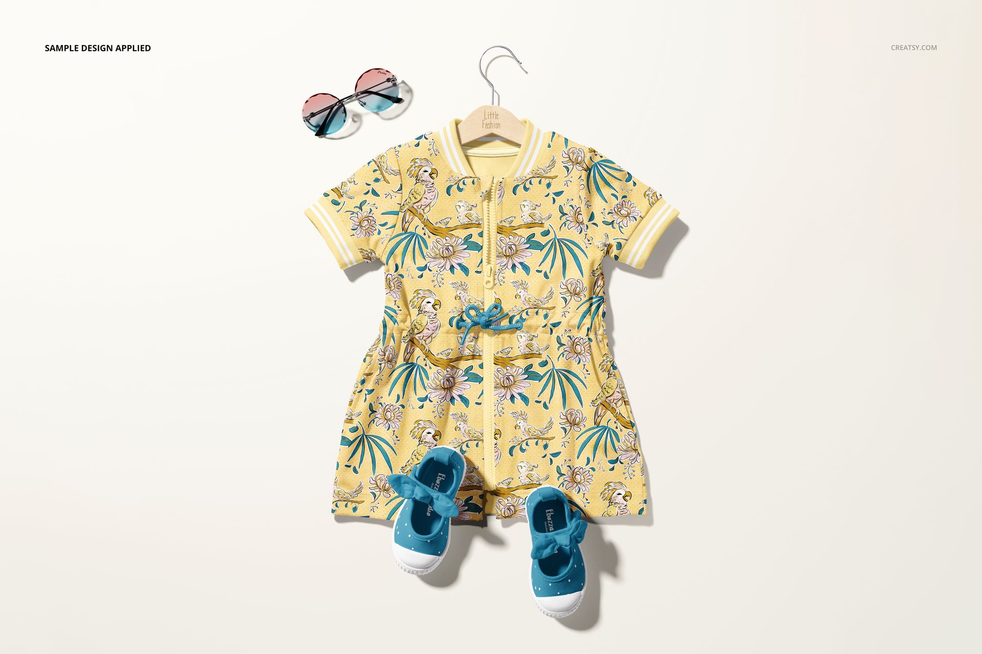 Front view of a baby dress mockup with a yellow tropical pattern, displayed on a hanger with blue shoes and sunglasses.