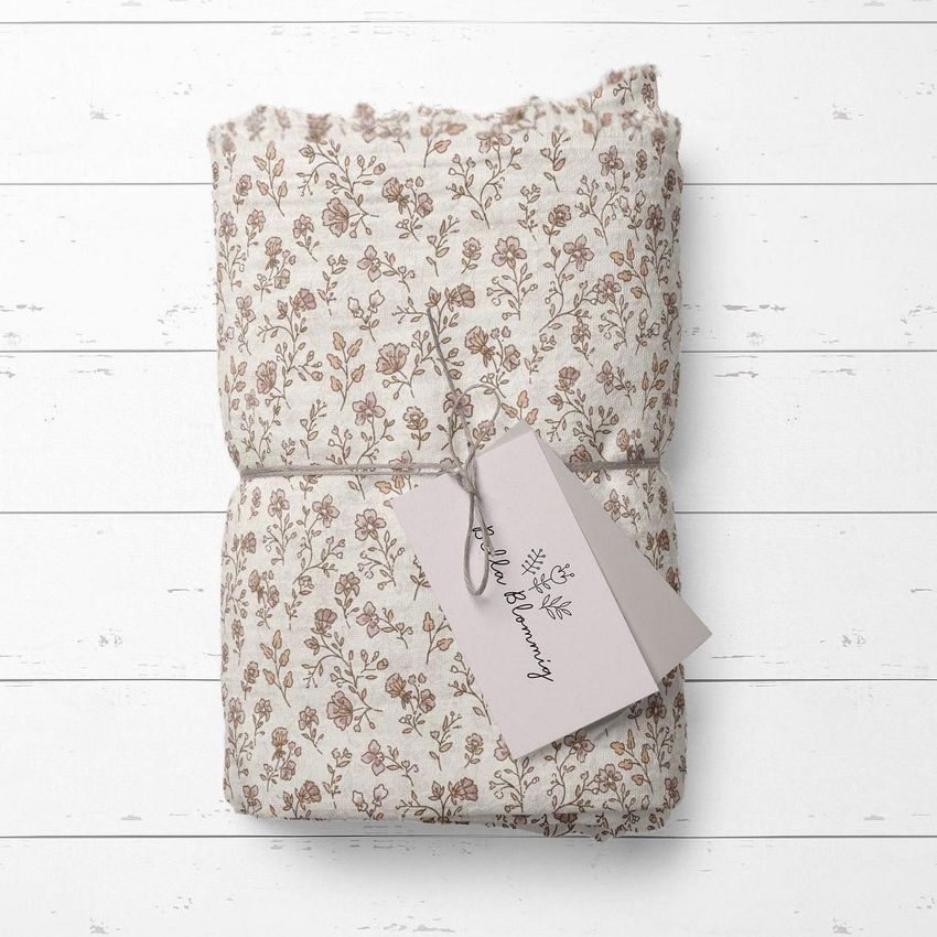 Muslin swaddle blanket mockup featuring a folded fabric with a delicate floral pattern, tied with string and a blank tag on a white wooden surface.