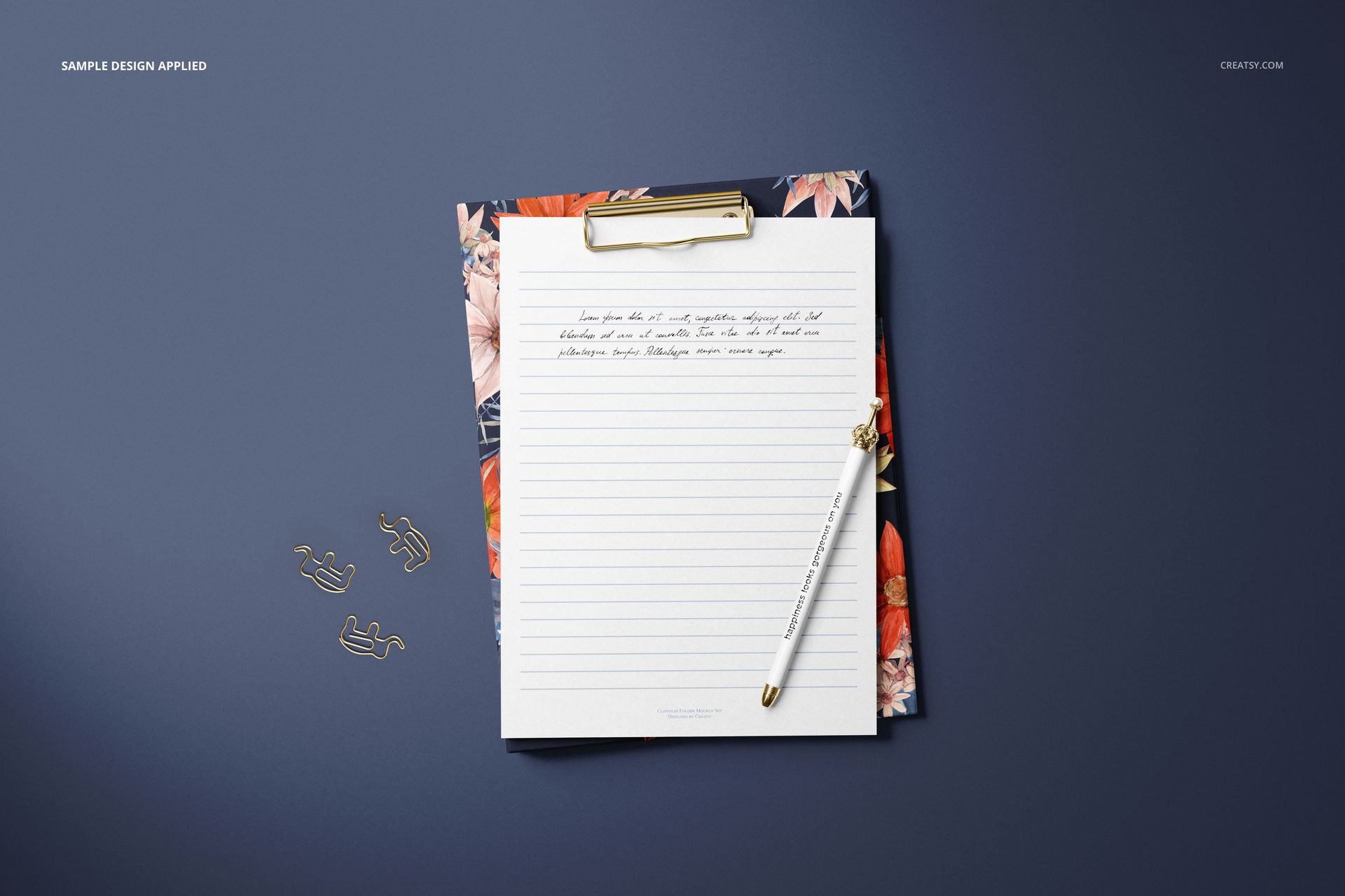 Stationery mockup featuring a lined paper sheet with handwritten text, gold clip, pen, and floral backing on a dark surface.