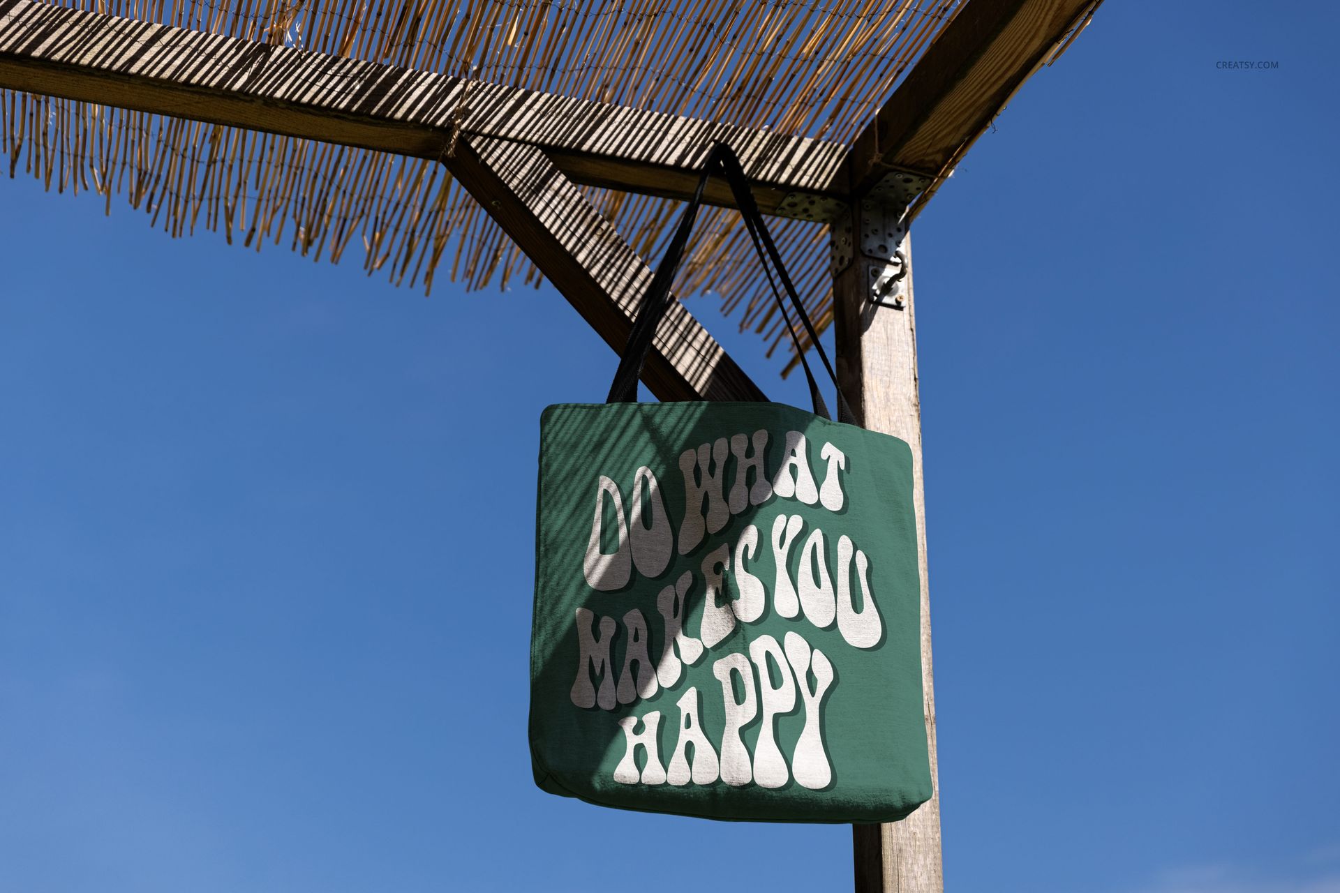 Woven tote bag mockup featuring a green bag with white text, hanging beneath a bamboo roof against blue sky.