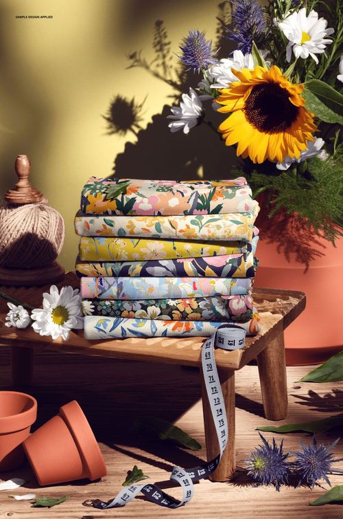 PSD mockup showing folded jersey fabrics with various flower prints, set on a wooden stool with garden-themed props.