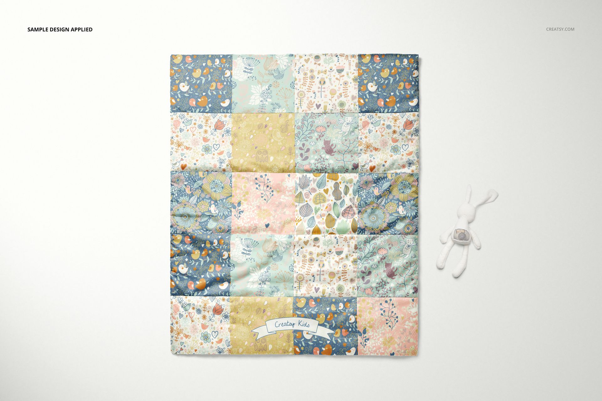 Top view of a baby patchwork quilt mockup featuring soft blue, yellow, and pink panels in floral designs beside a plush rabbit.