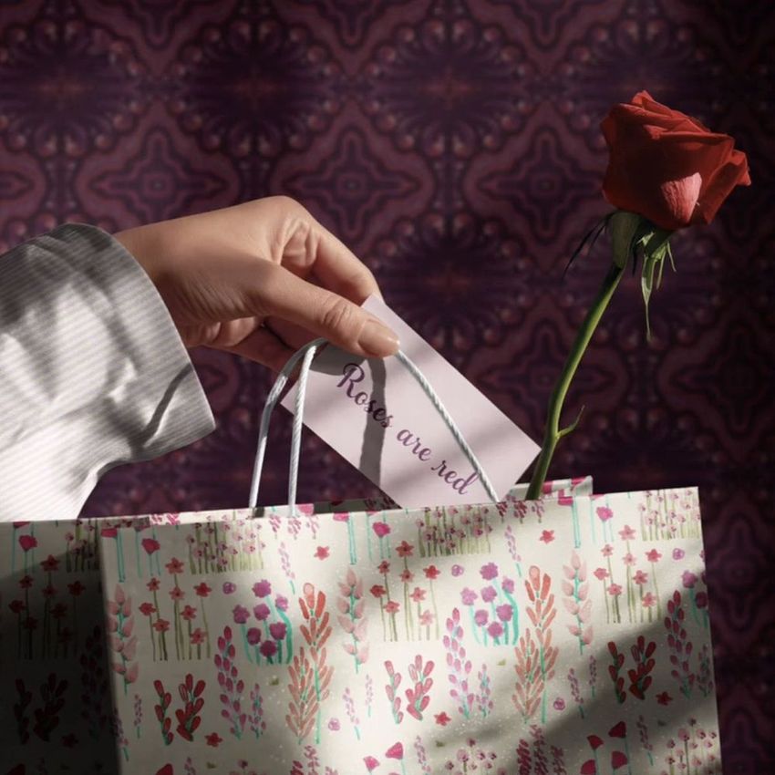 Hand inserting a card with visible text into a floral gift bag mockup, with a single red rose inside, against a patterned background.
