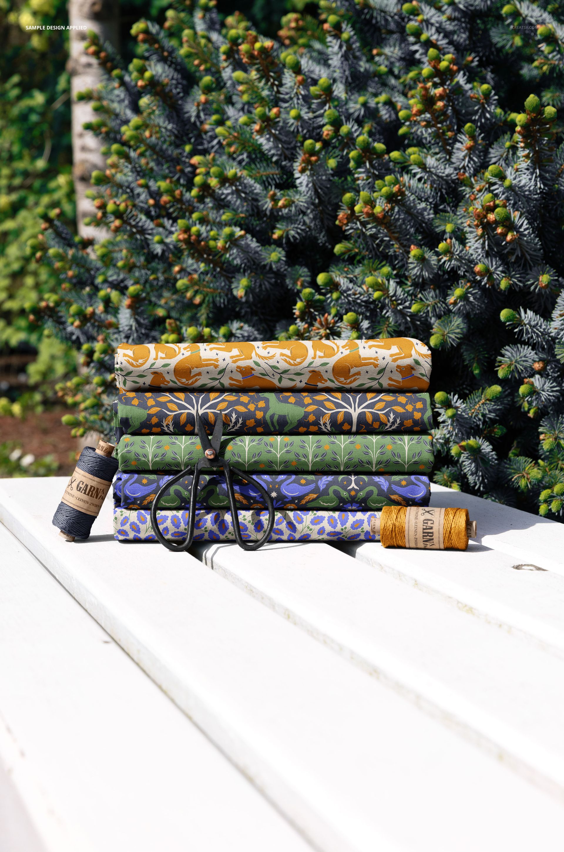 PSD mockup showing colorful patterned fabrics tied with string, placed on a white bench near evergreen trees.