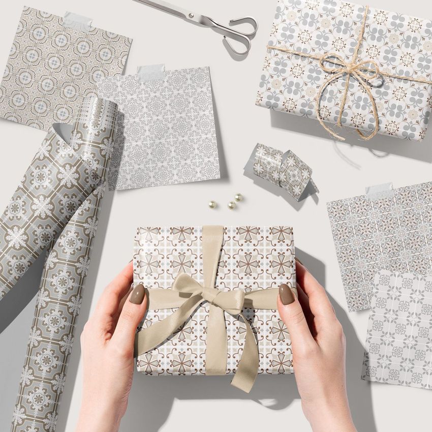 Gift wrapping paper mockup featuring hands holding a wrapped box with beige ribbon, surrounded by patterned paper sheets and scissors.