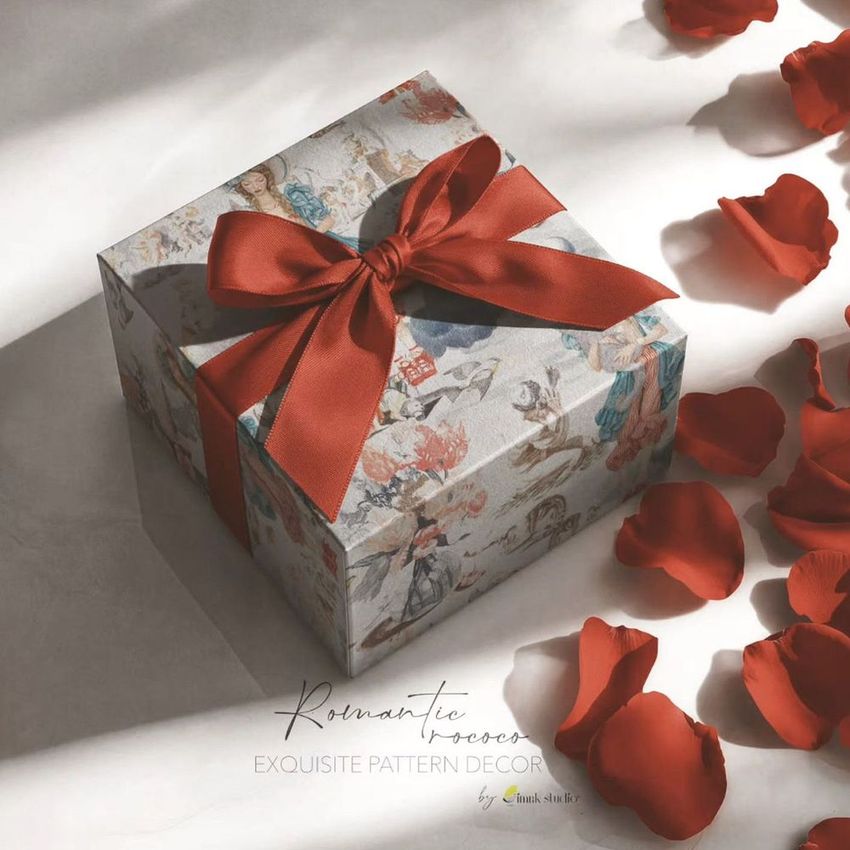 Square gift box mockup with a red ribbon bow, decorated with a rococo-style pattern, surrounded by scattered red rose petals.