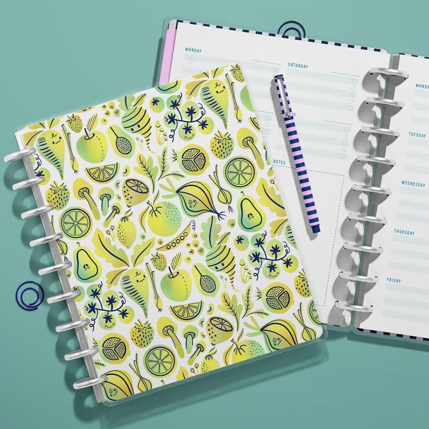 Top view of two spiral planners on a teal surface, one closed with a colorful fruit pattern cover, the other open to a weekly layout.