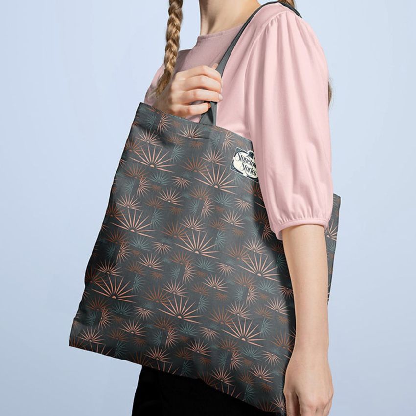 Tote bag mockup featuring a woman in a pink blouse holding a dark patterned tote with geometric sunburst designs.
