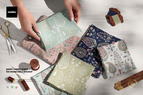 PSD mockup showing several folded jersey fabric swatches in pastel and dark tones, surrounded by sewing tools.