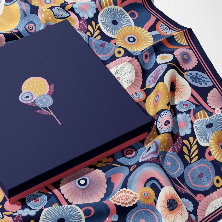 Silk scarf box mockup featuring a navy blue box with a floral graphic, placed on a colorful patterned silk scarf.