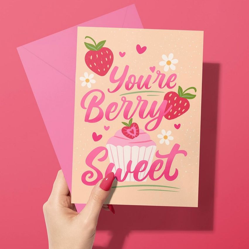 Greeting card mockup featuring a woman's hand holding a card with strawberries, flowers, and cupcake illustration on a pink background.
