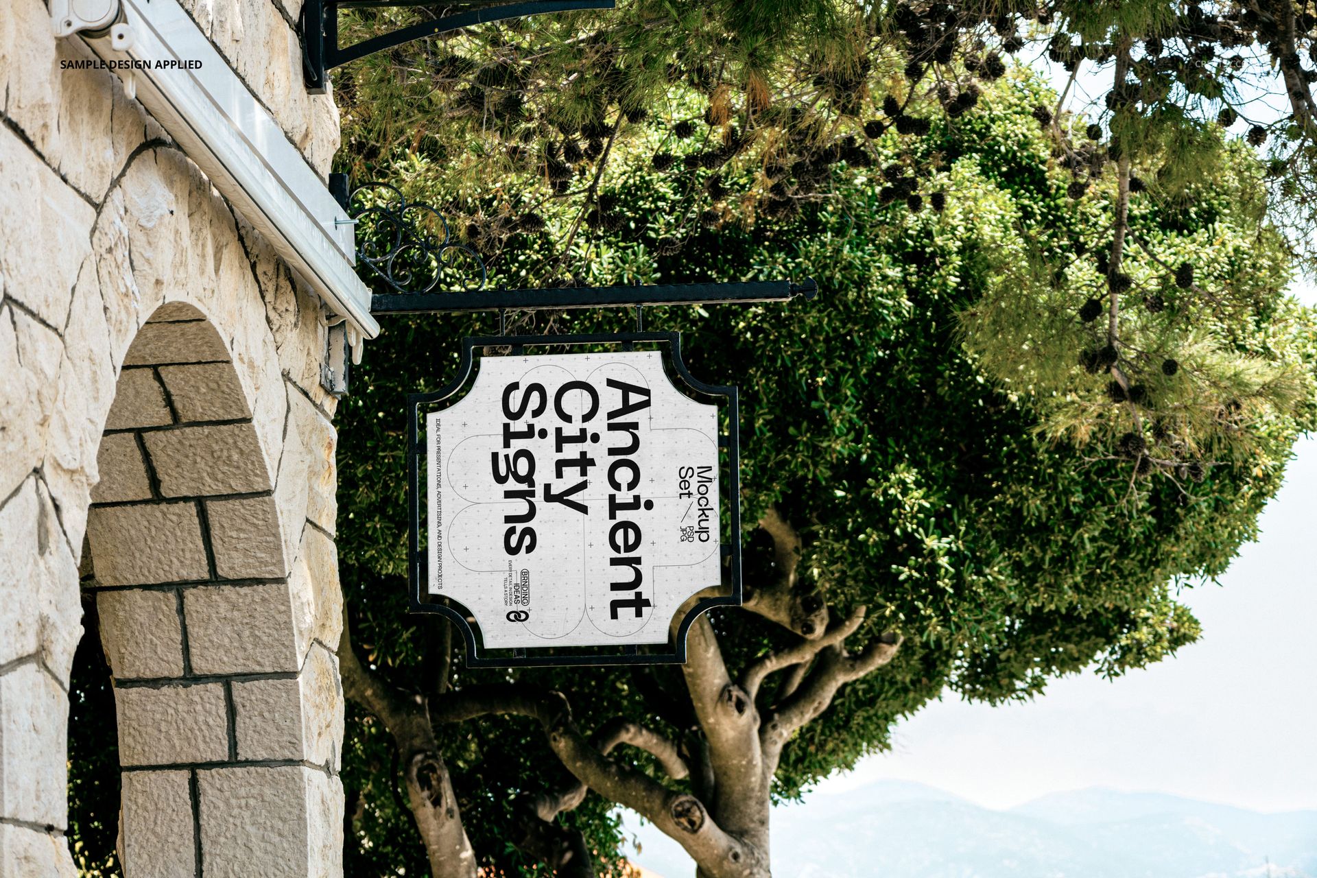 Rectangular city sign mockup hanging on a decorative metal bracket, stone building wall and green trees in background.