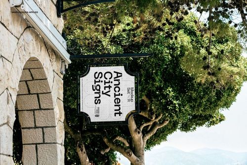 Rectangular city sign mockup hanging on a decorative metal bracket, stone building wall and green trees in background.