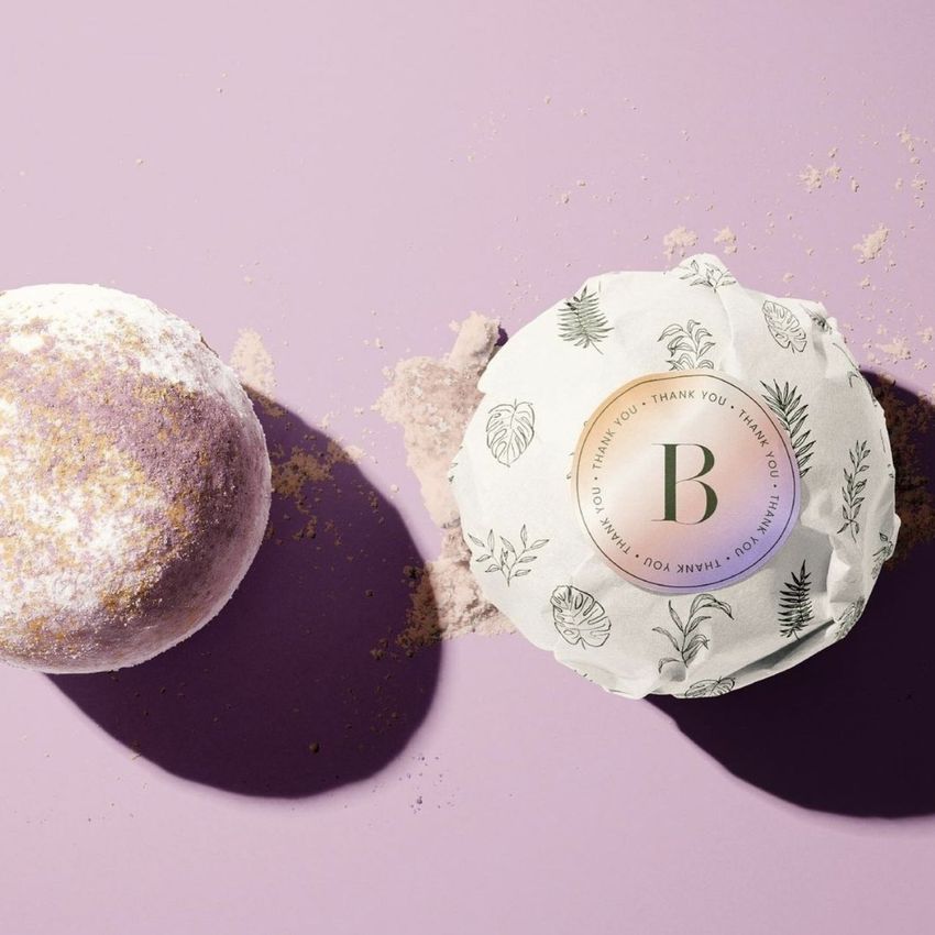 Sticker mockup featuring a round package wrapped in white paper with a botanical pattern and a circular sticker on top.