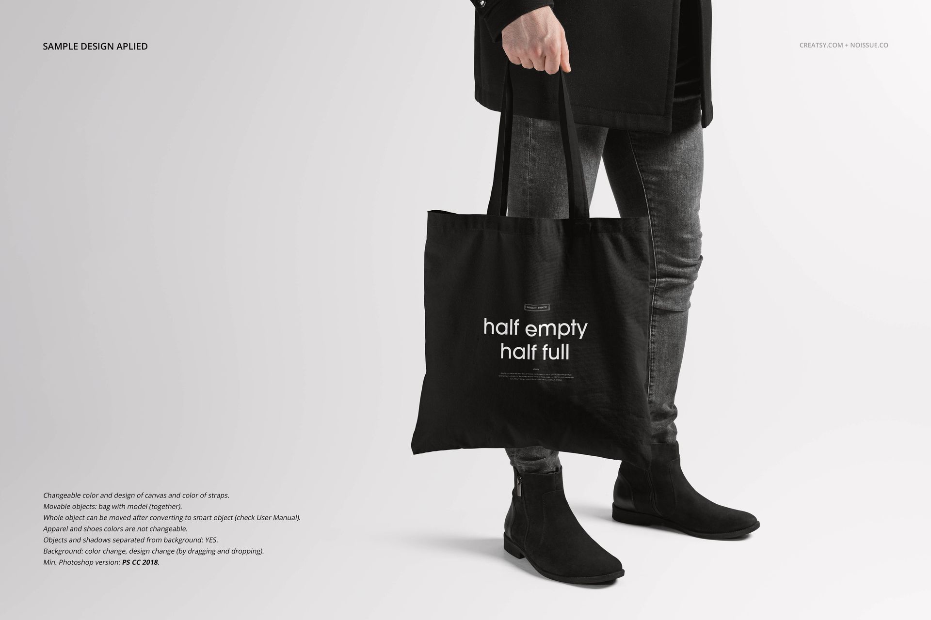 Front view of a black tote bag mockup held by a person wearing dark clothing and boots against a light background.