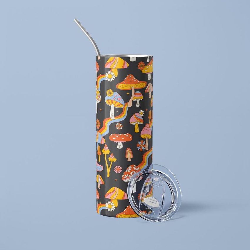 Skinny tumbler mockup with a colorful mushroom and rainbow pattern on a dark background, shown upright with a metal straw and a clear lid beside it on a light blue surface.