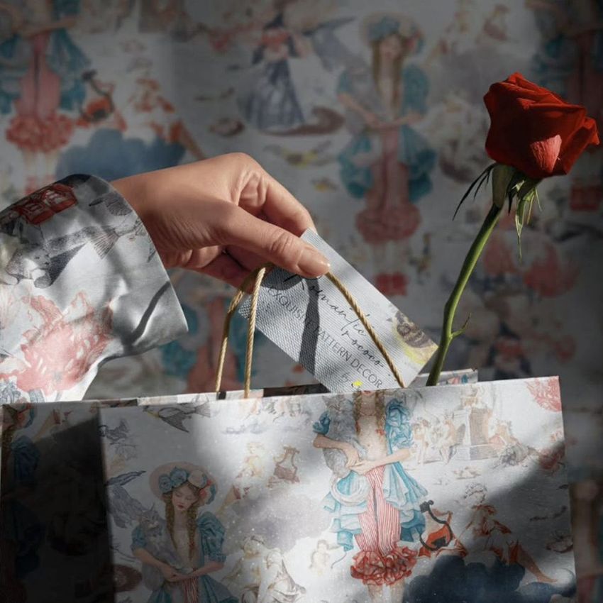 Hand inserting a card into a patterned gift bag with a single red rose, set against a matching decorative background.