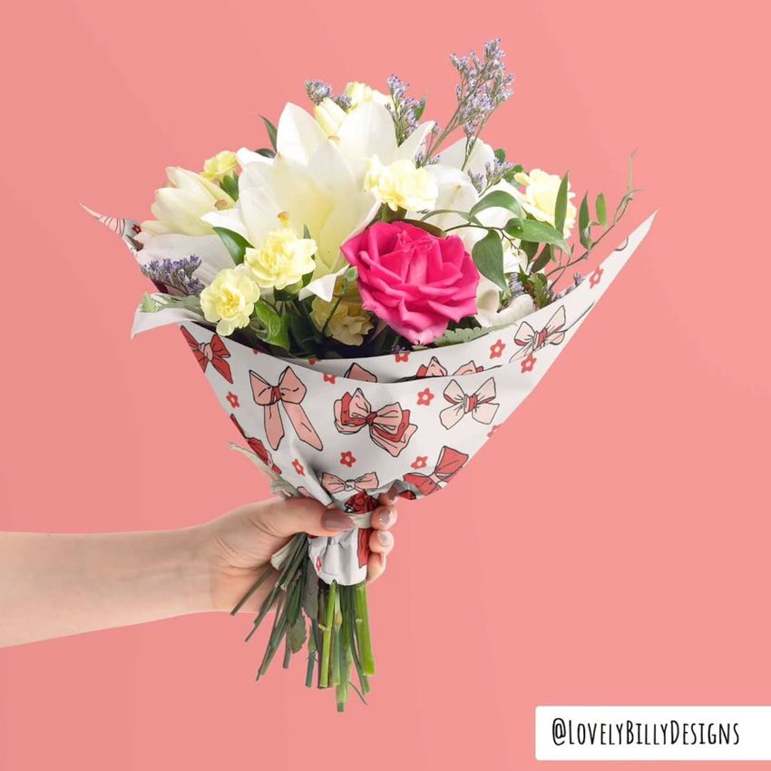 A hand holds a bouquet of assorted flowers wrapped in white packaging with red and black bow patterns, set against a pink background.
