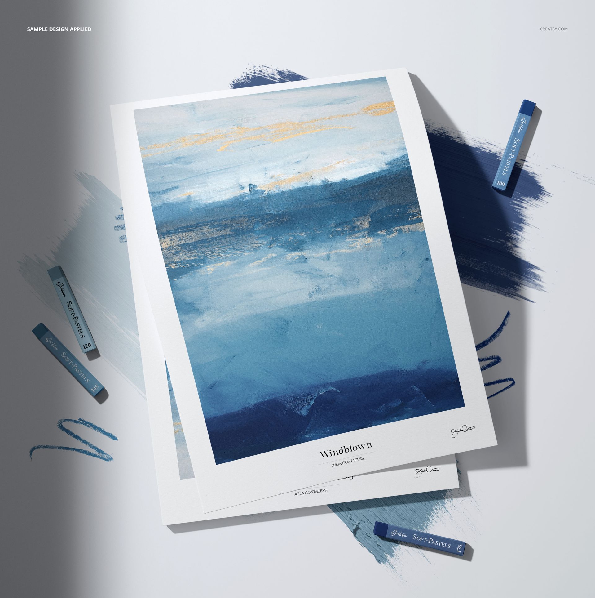 Soft pastel sticks in shades of blue arranged around decorative paper featuring an abstract blue artwork, top view.