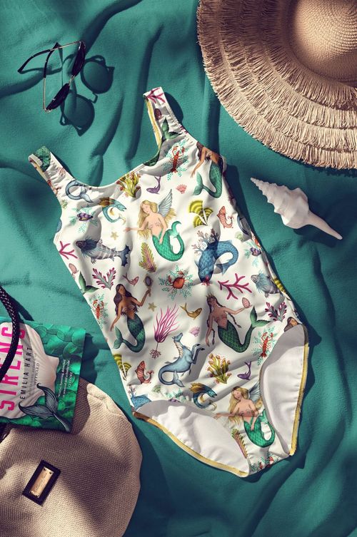 Swimsuit mockup with whimsical sea creature design, displayed on teal cloth alongside a book, bag, and summer props.