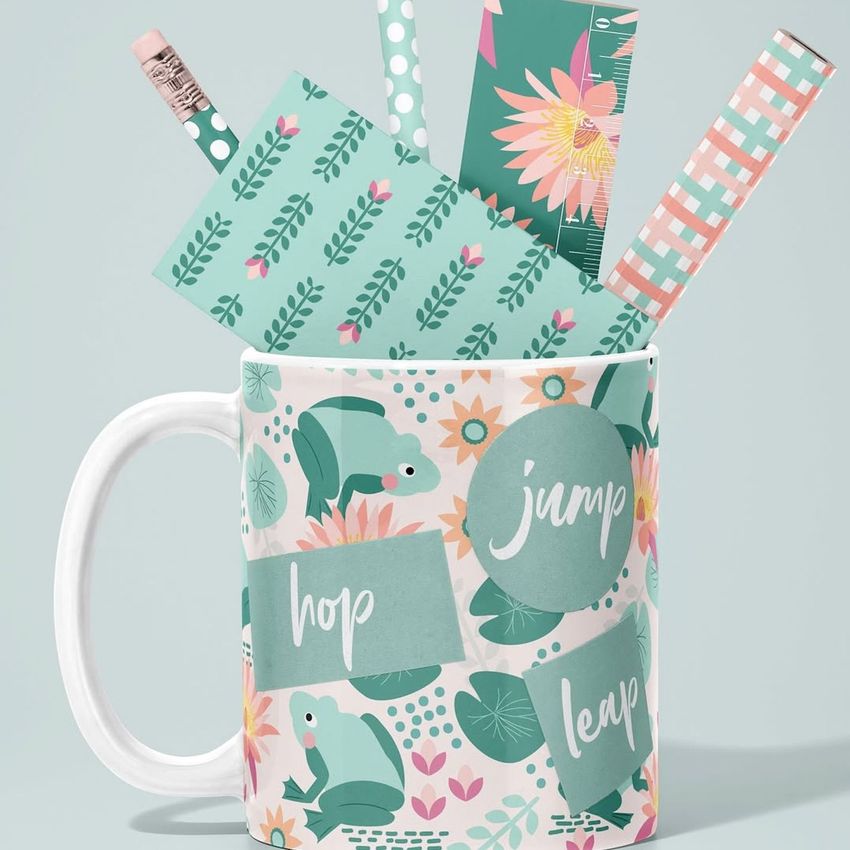 Mug mockup with illustrated frogs, flowers, and leaves in green and pink, filled with patterned pencils and bookmarks.