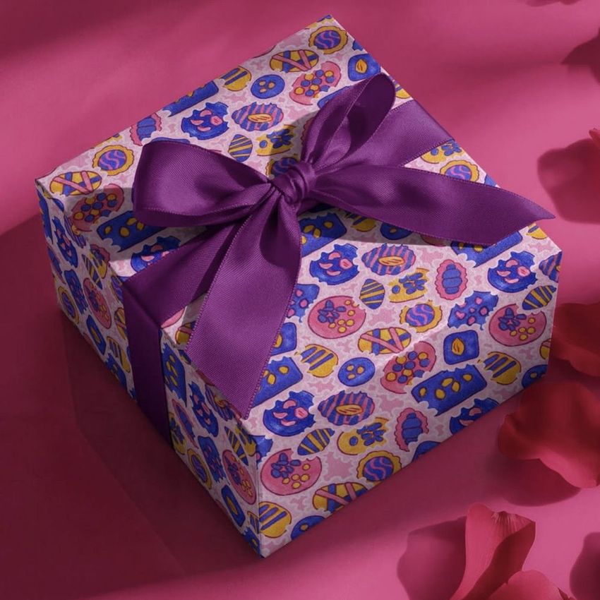 Square gift box mockup with a purple ribbon, decorated with colorful abstract patterns, placed on a pink surface with scattered petals.