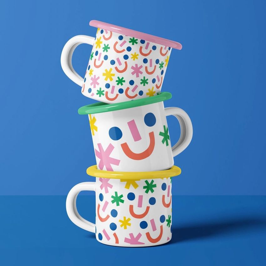 Three enamel mug mockups with colorful, playful smiley face patterns are stacked against a solid blue background.