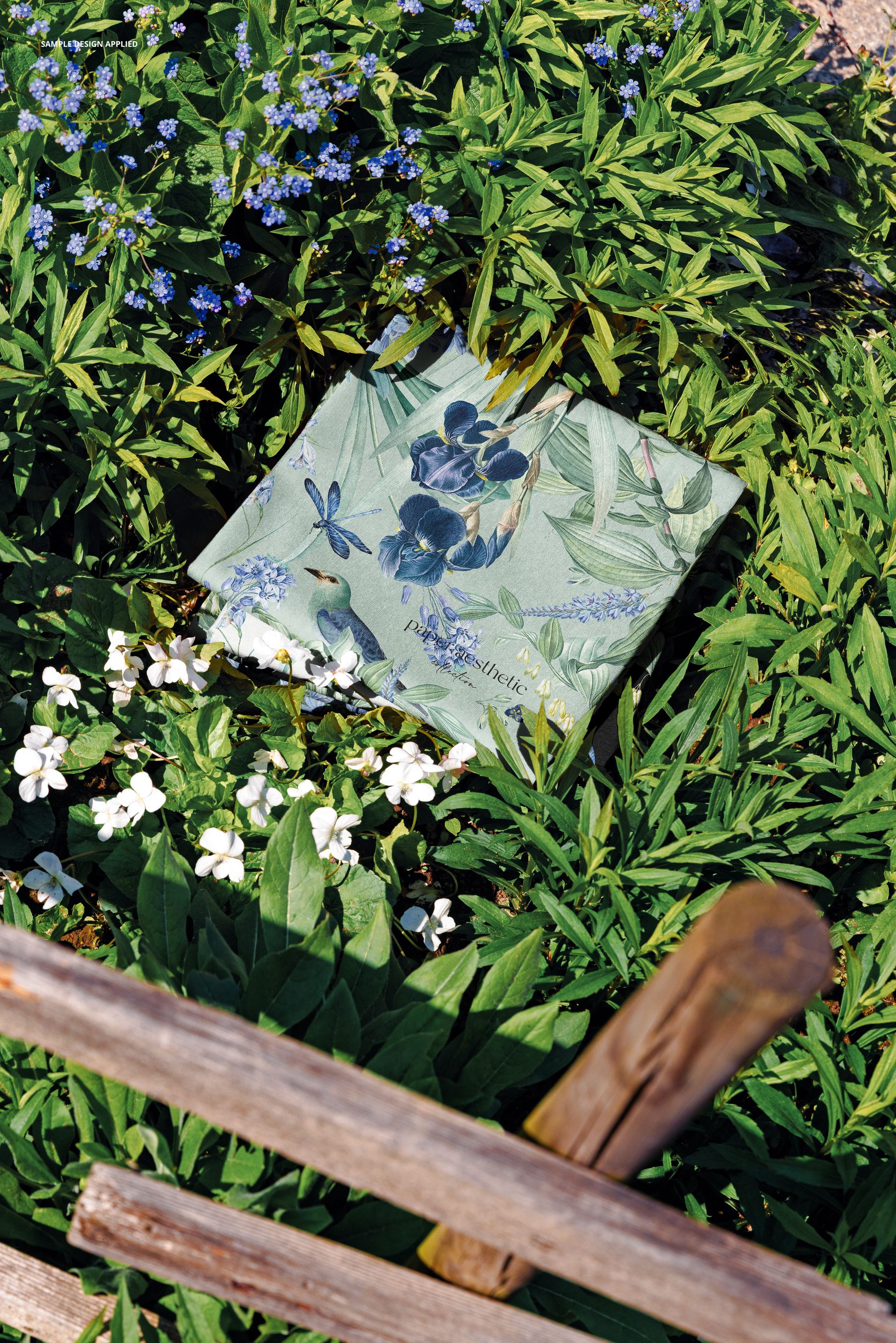 PSD mockup of patterned fabric with blue and green floral motifs lying on a leafy garden ground with white blossoms.