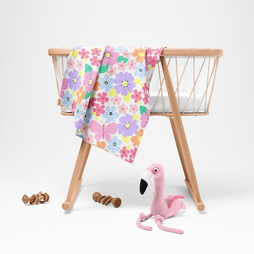 Crinkle muslin swaddle blanket mockup draped over a wooden baby bassinet, featuring a colorful floral pattern and a pink plush flamingo toy on the floor.