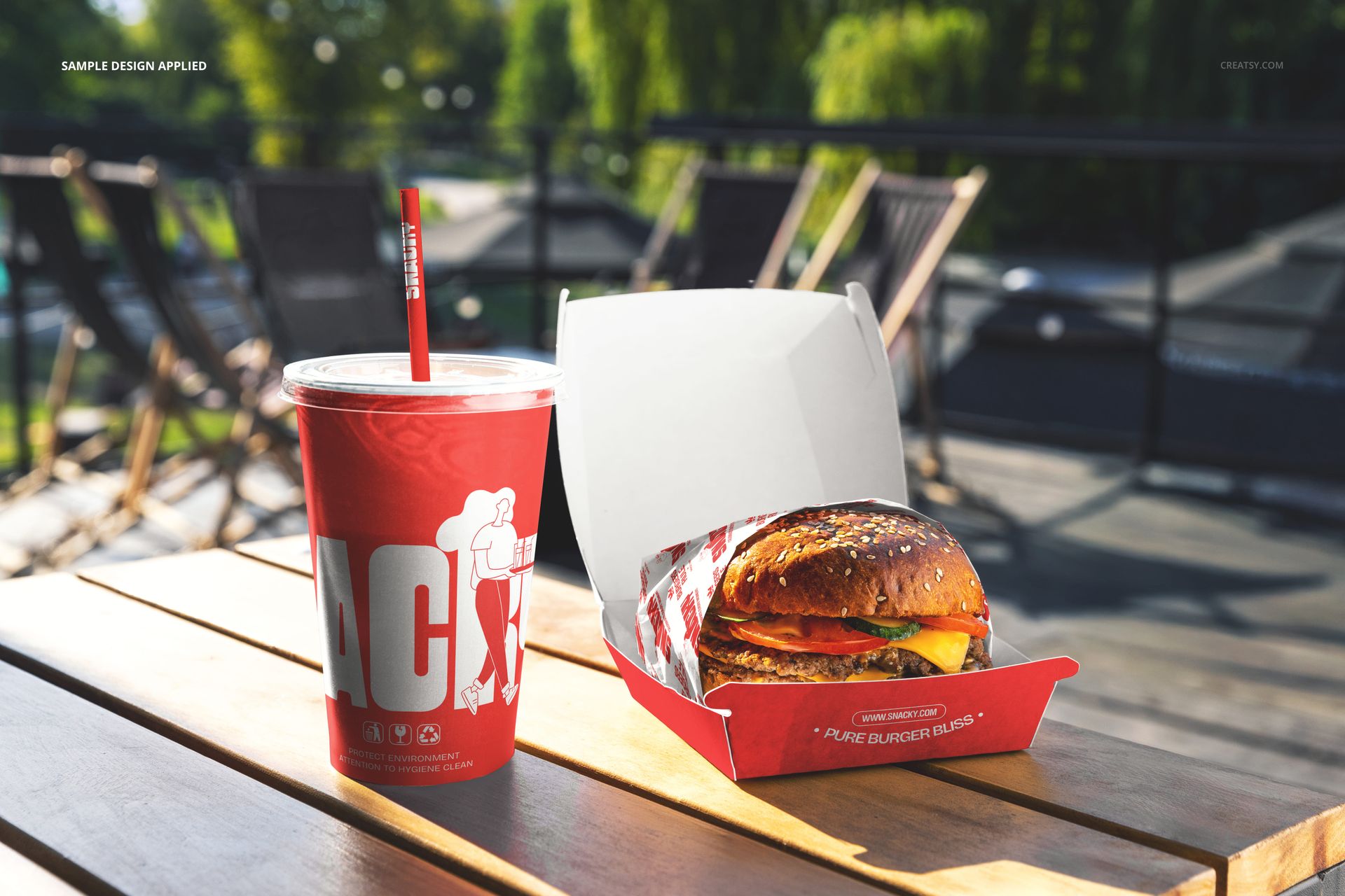 PSD mockup showing a burger and red beverage cup with straw on a picnic table, surrounded by outdoor seating and greenery.