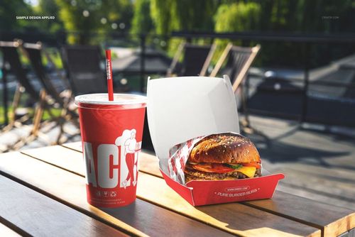 PSD mockup showing a burger and red beverage cup with straw on a picnic table, surrounded by outdoor seating and greenery.